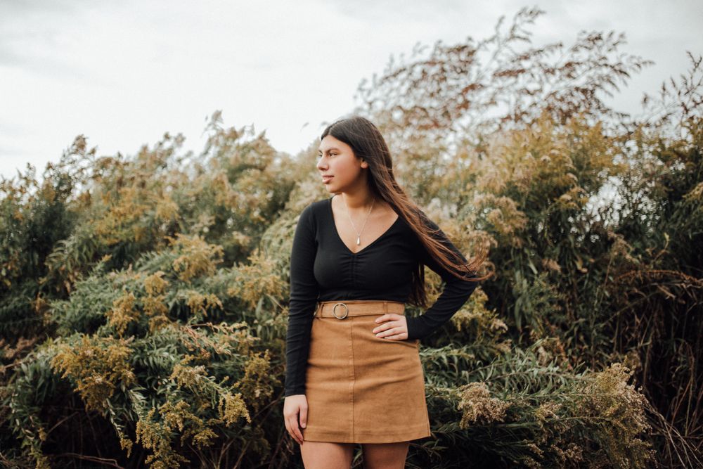 Person in black top and brown skirt posing against windswept natural backdrop.