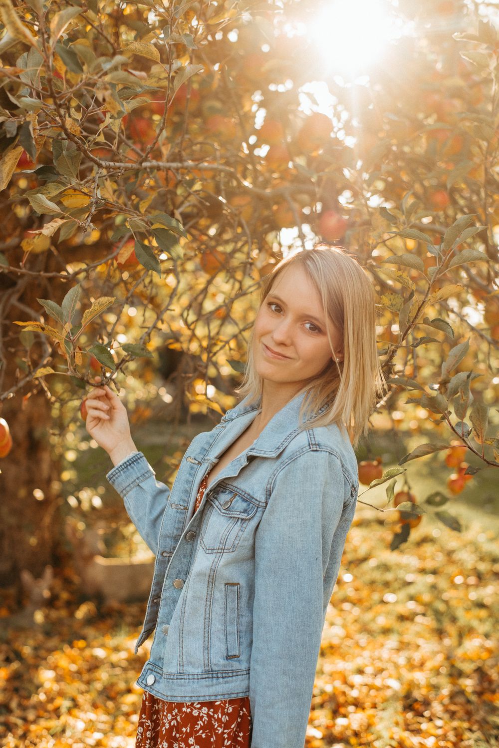 Woman in denim jacket poses among golden autumn leaves and sunlight.