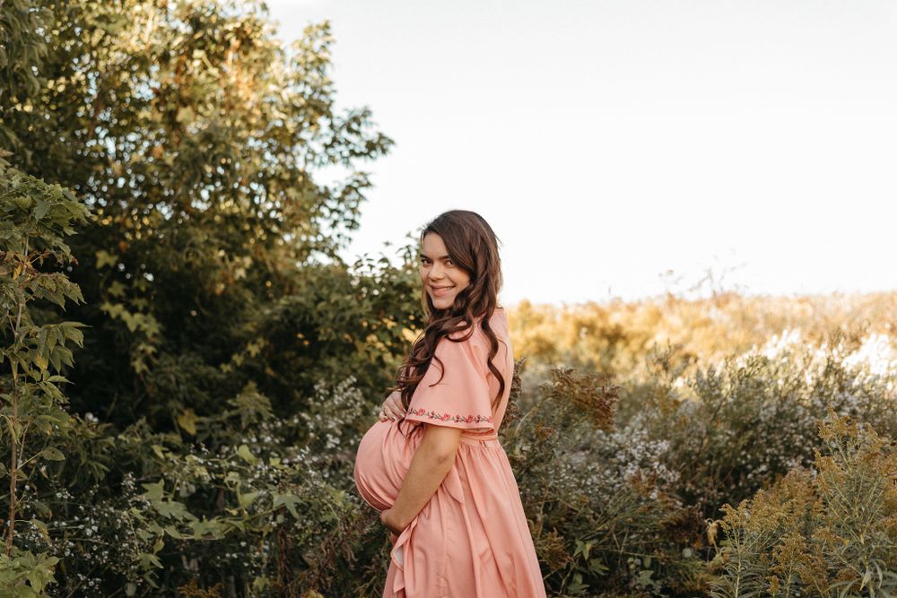 Expecting mother in pink dress poses in natural setting during golden hour.