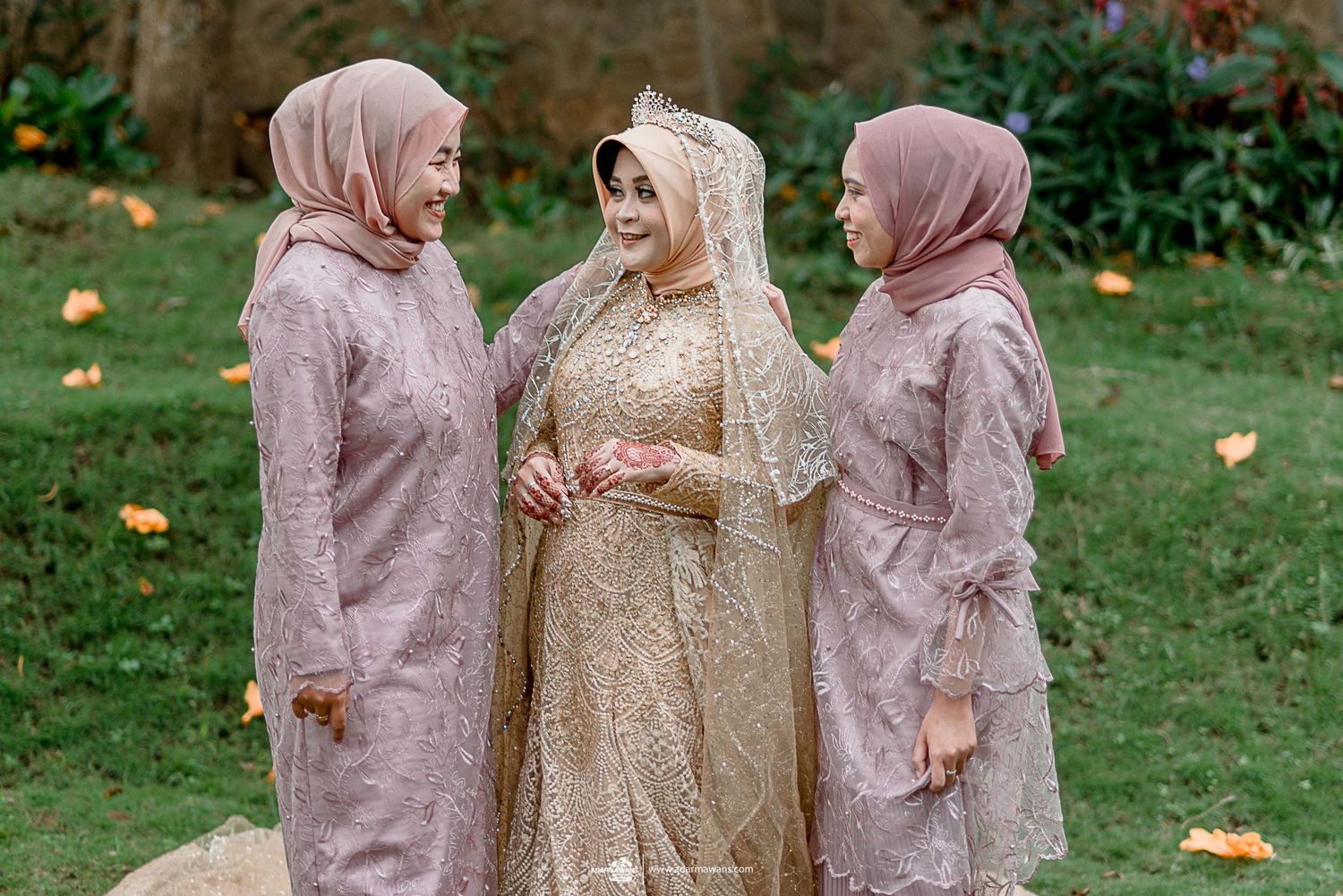 Three women in coordinating dusty rose and gold dresses share a joyful moment outdoors.
