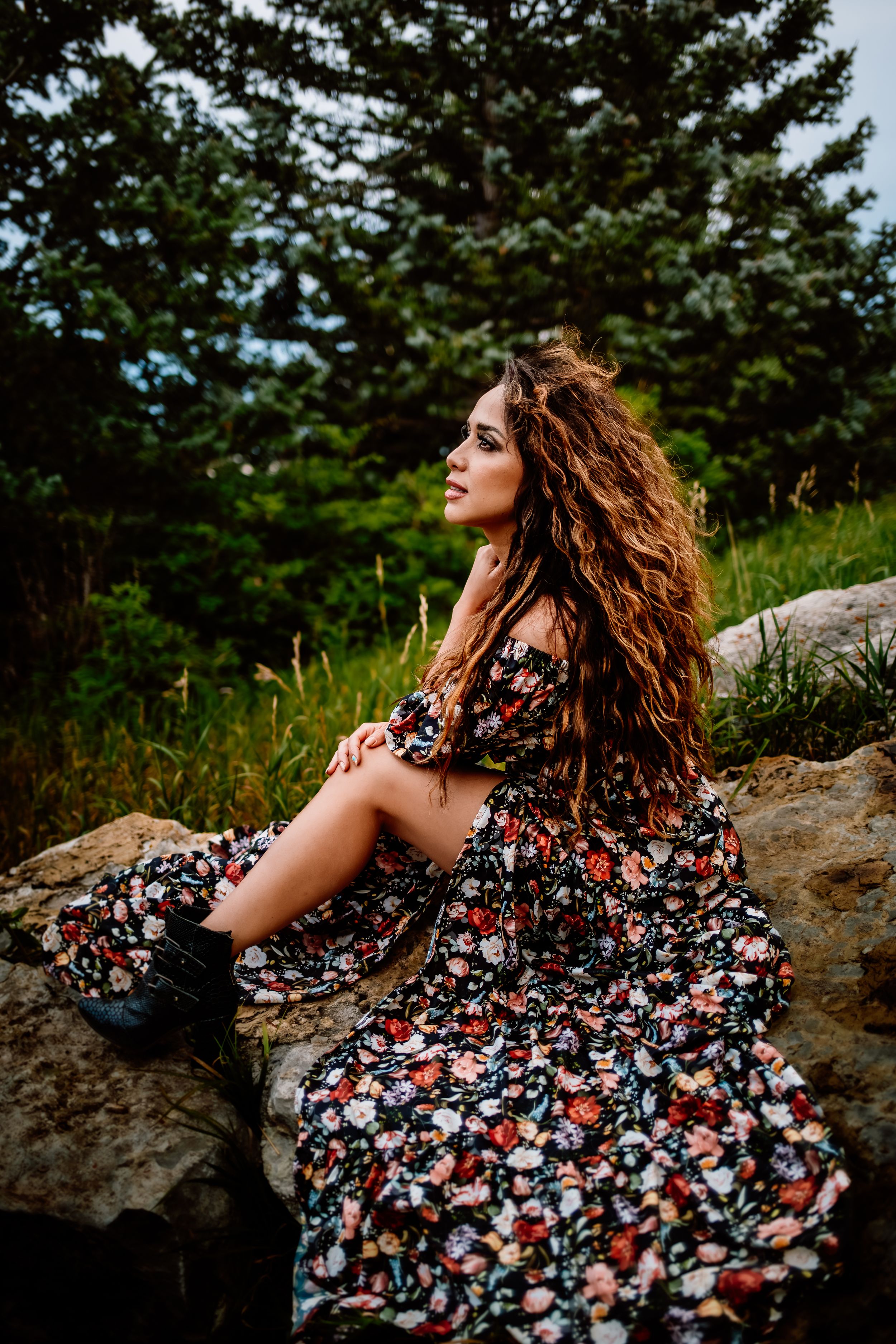 Woman in floral dress poses on rocky terrain with evergreen trees in background.