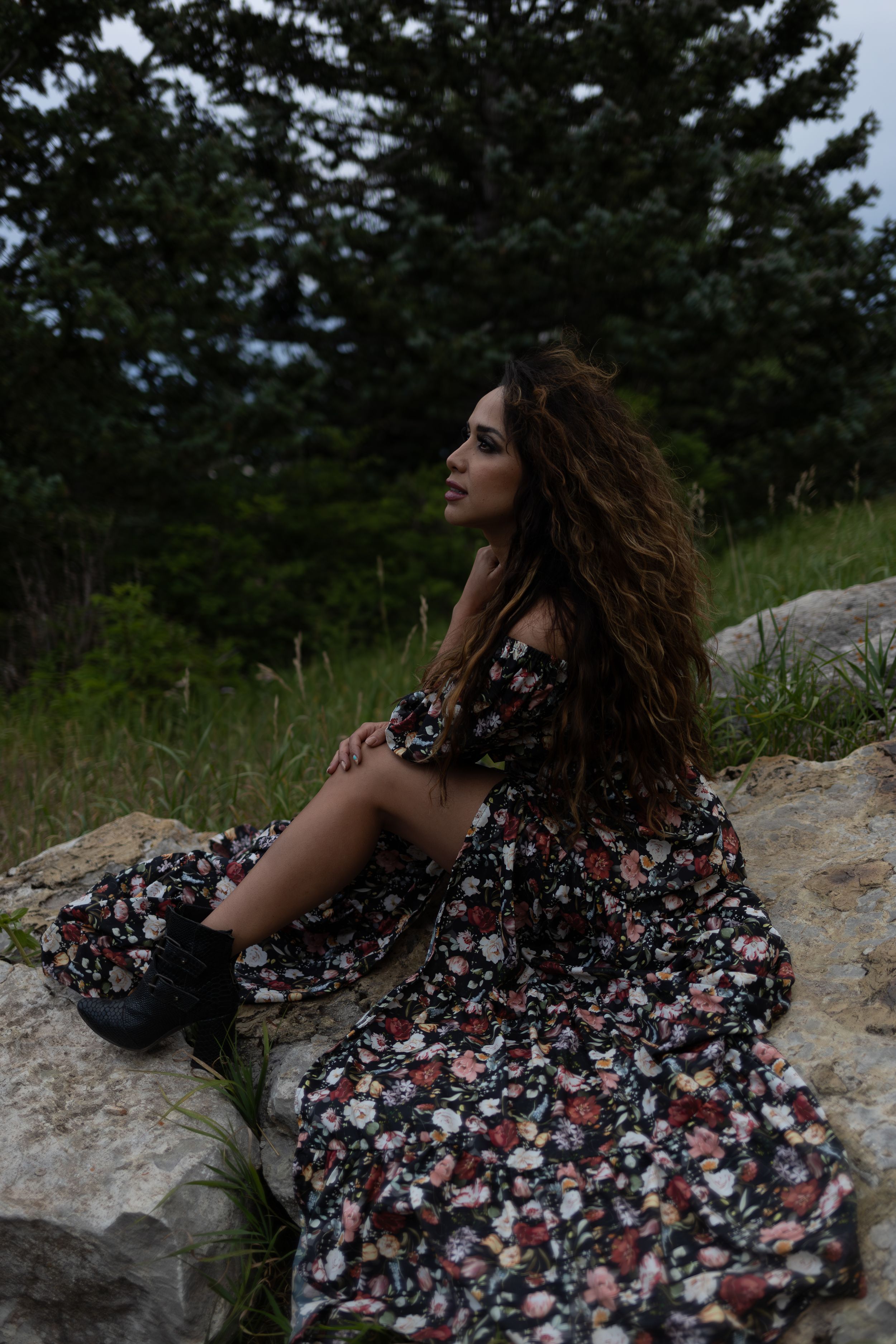 Moody portrait of woman in floral dress sitting on rocky outcrop near pine trees.
