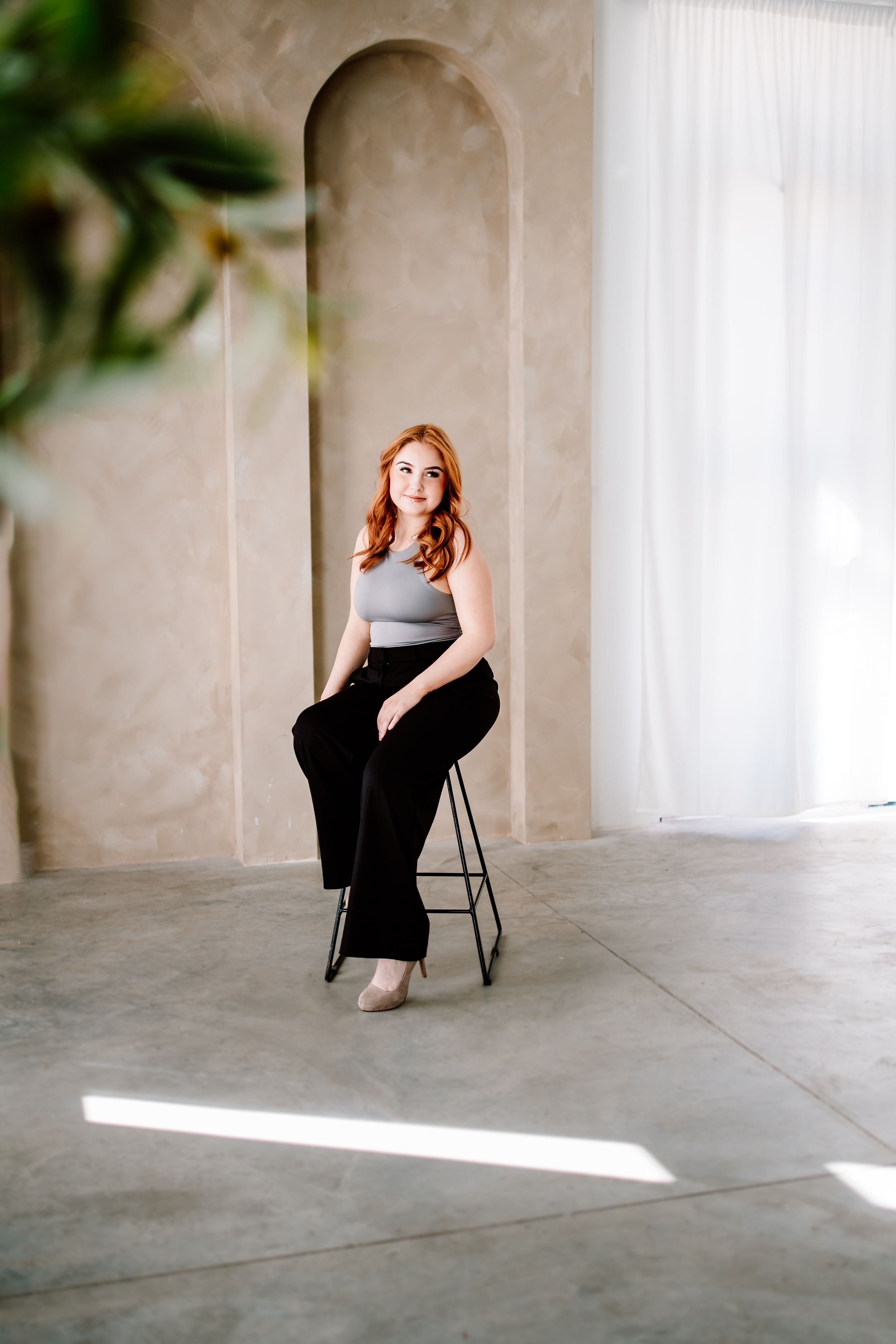A studio portrait featuring a seated figure in a grey top and black pants against a minimalist architectural backdrop.