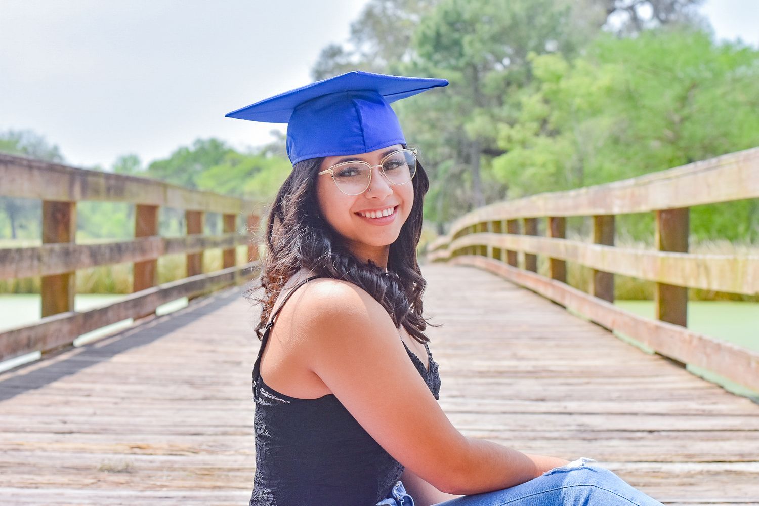 Graduate in blue cap and gown poses on wooden boardwalk during graduation photoshoot.