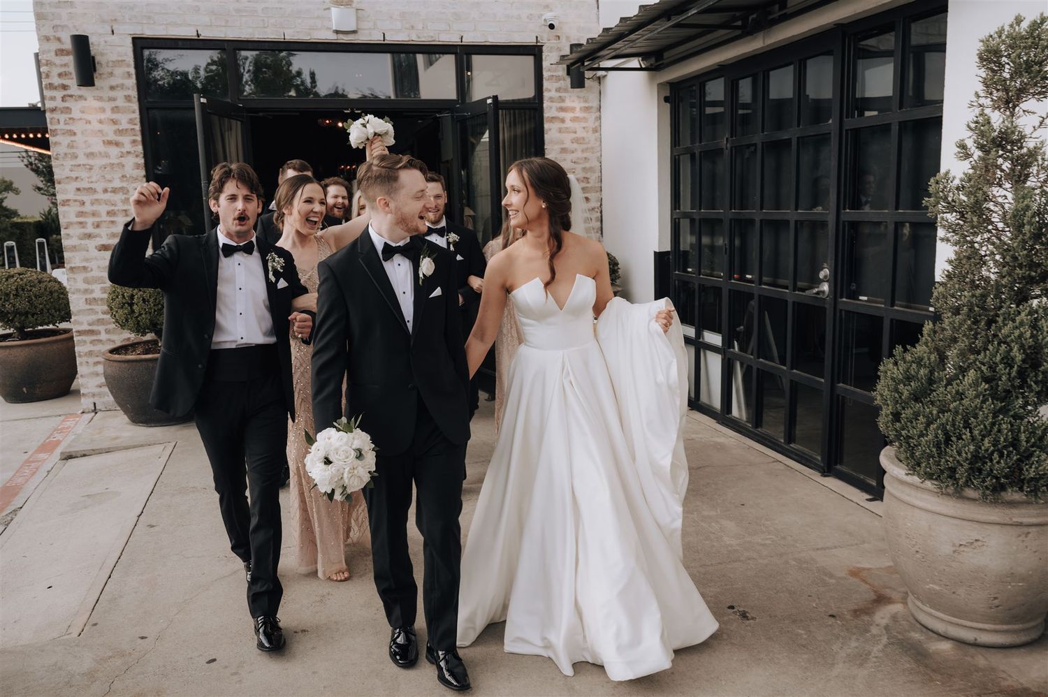 Joyful wedding party walking outside modern building with brick walls.