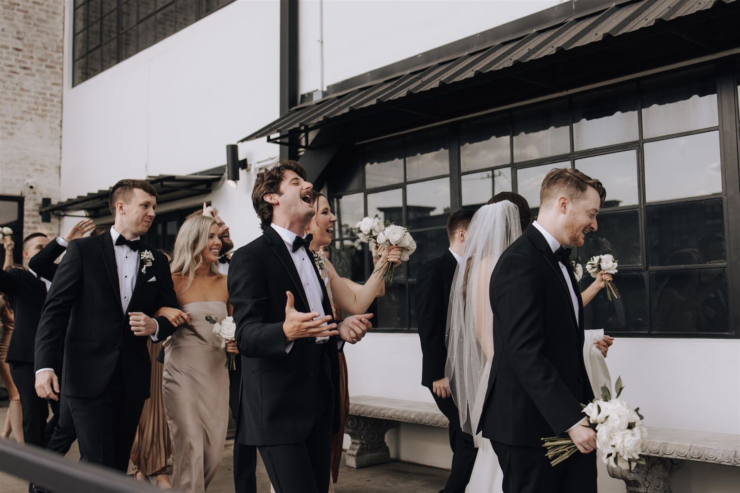 Wedding party members in formal attire walking along building exterior.