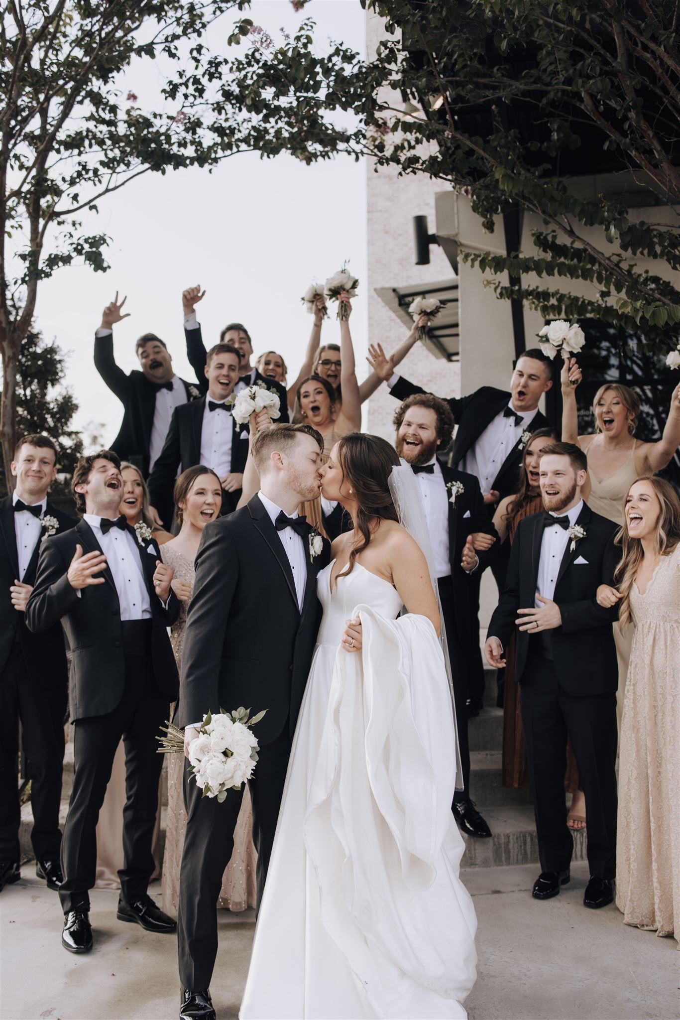 Wedding party cheers and celebrates with newlyweds outdoors in formal attire.