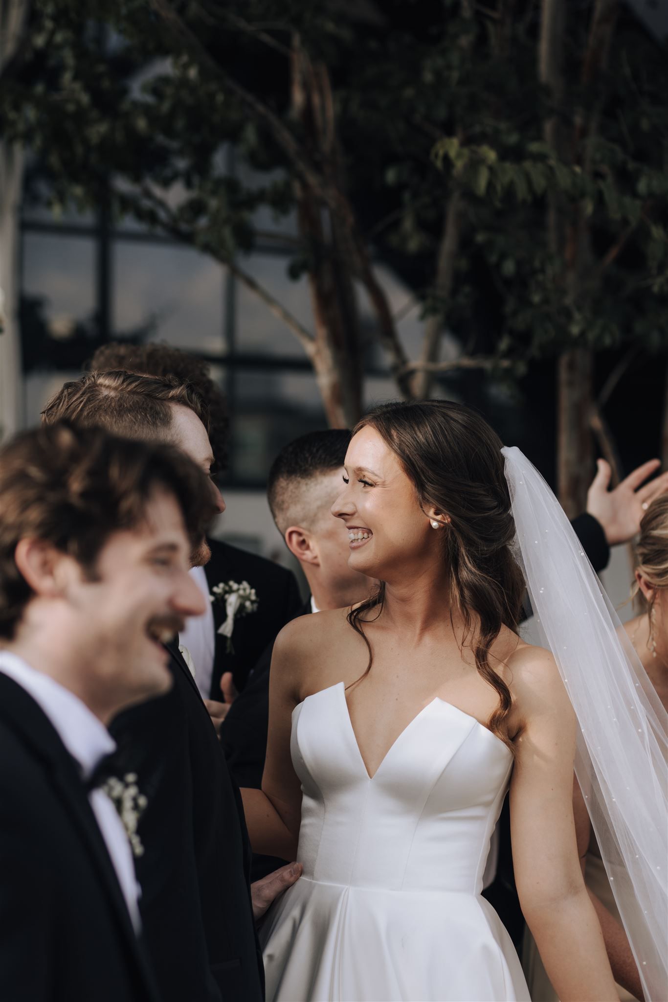 Wedding party laughing together outdoors with bride in elegant white dress and groom in black tuxedo.