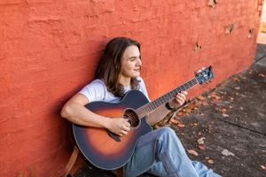 Musician playing acoustic guitar while sitting against vibrant orange wall.