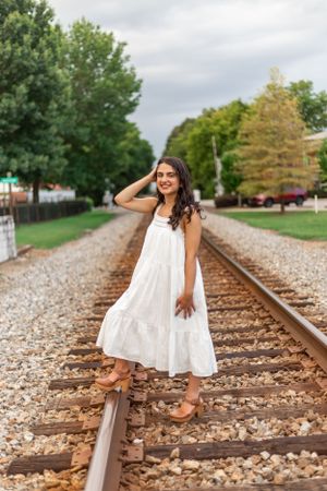 Person in white sundress poses playfully on train tracks surrounded by trees.