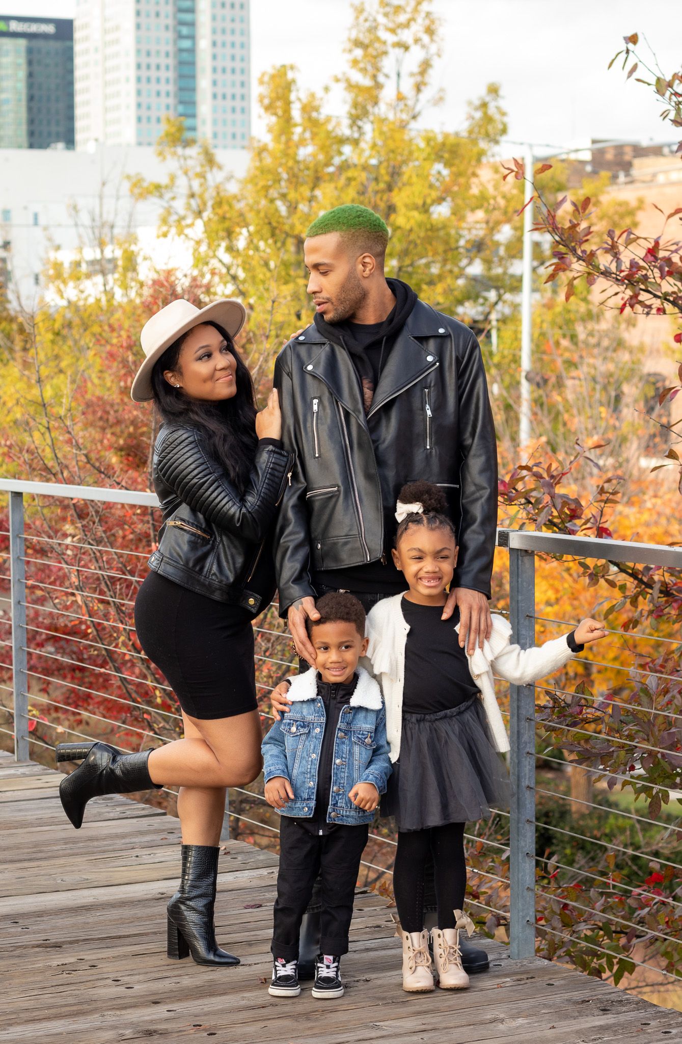 Stylish family of four in autumn outfits posing on wooden deck with city backdrop.
