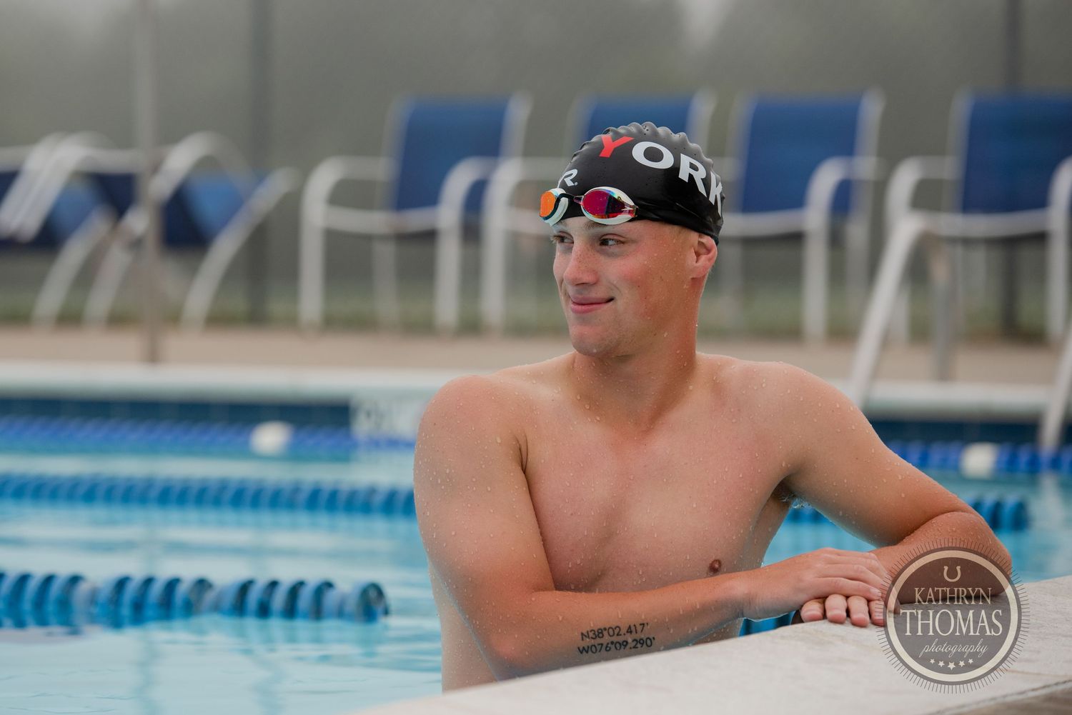 A swimmer in swim cap and goggles rests at the edge of a pool with blue chairs in background.