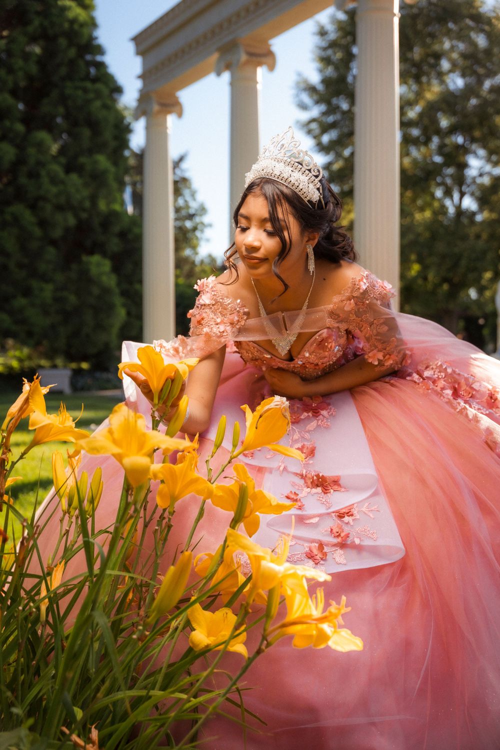 Young woman in pink floral ball gown and tiara admiring yellow lilies in a garden with white columns.