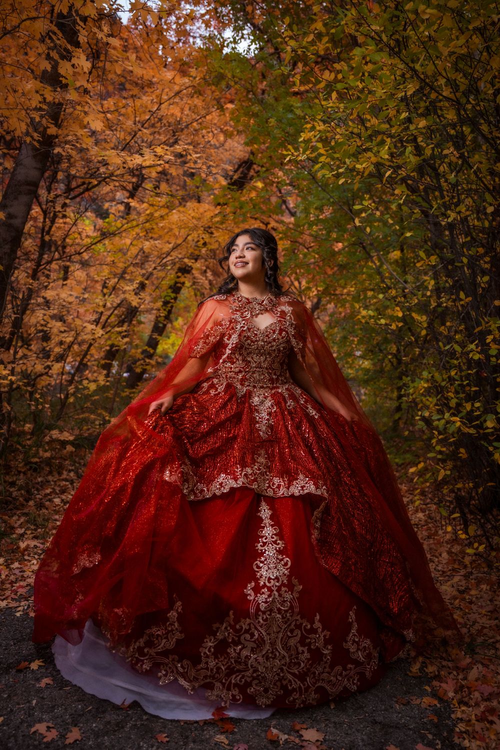 Young woman in a red sequined quinceañera gown with cape standing in an autumn forest setting.