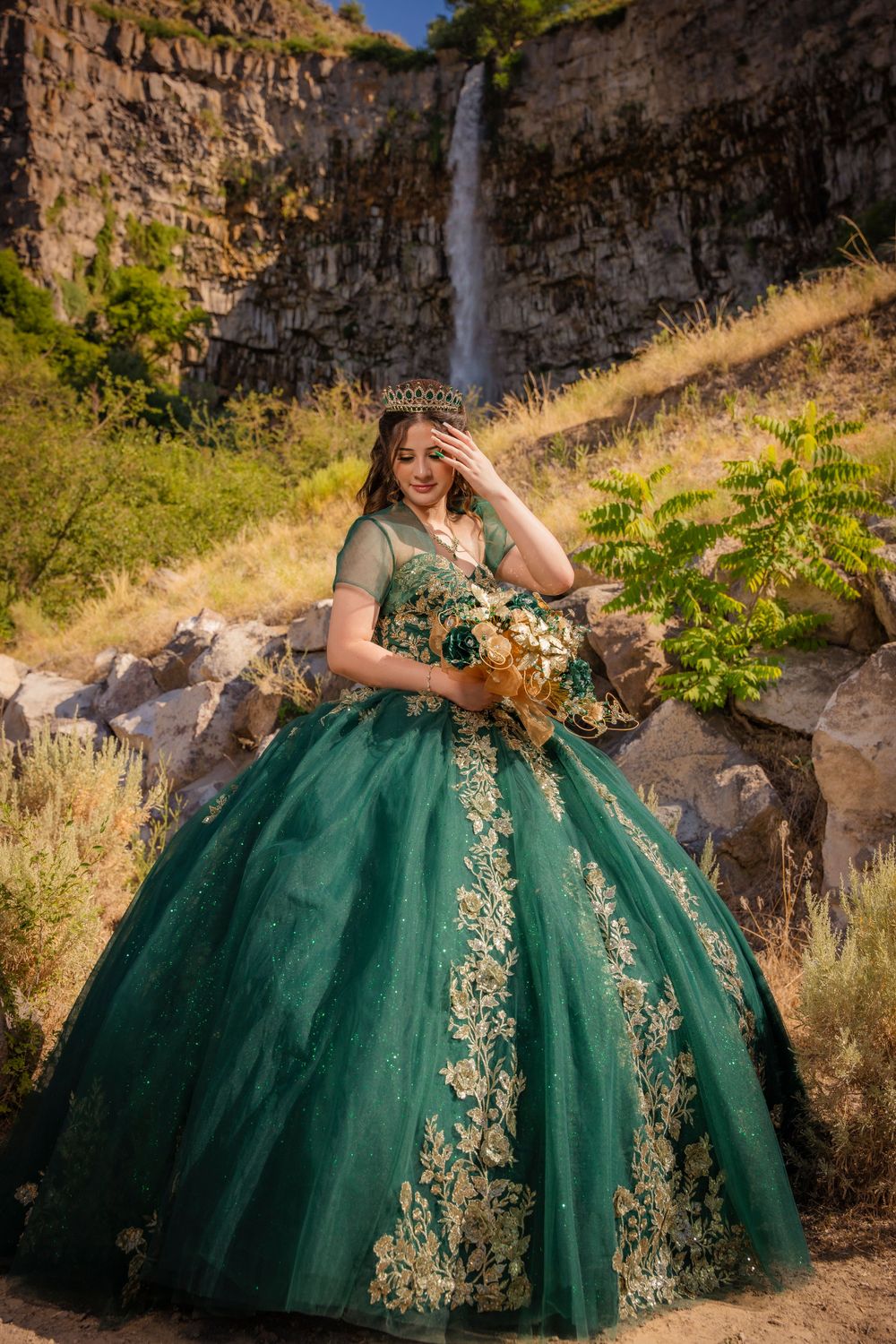 Young woman in emerald green quinceañera ballgown holding bouquet near a waterfall backdrop.