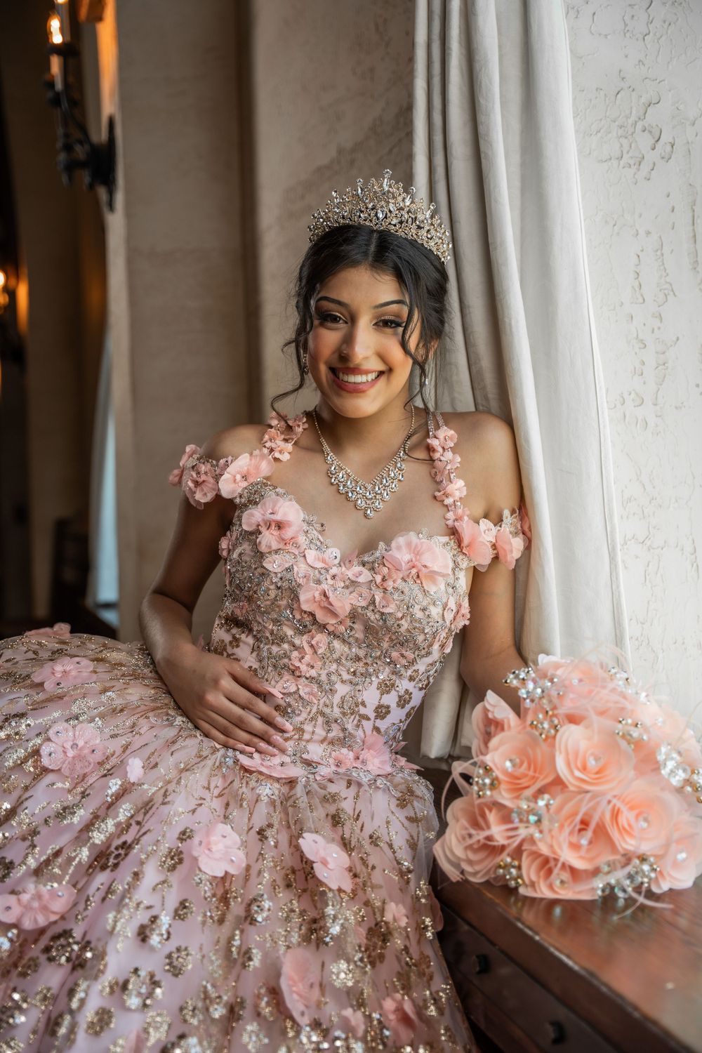 Smiling young woman in pink floral quinceañera gown with tiara and rose bouquet seated indoors.