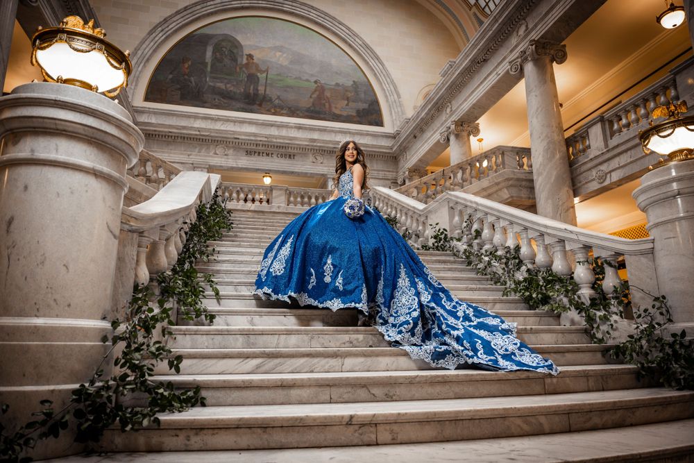 Elegant woman in a blue ball gown with white embroidery posing on grand marble staircase indoors.
