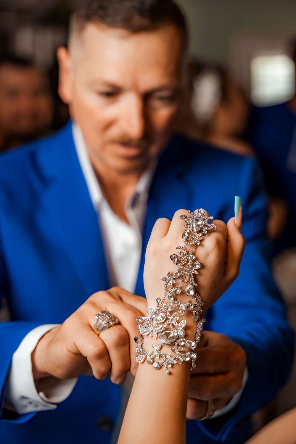 Man in blue suit carefully placing an ornate diamond floral hand jewelry piece on a young womans wrist.