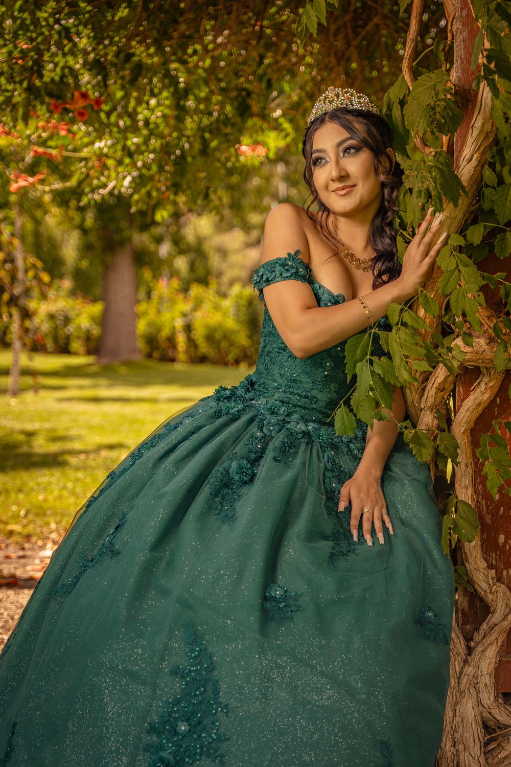 Young woman in an emerald green off-shoulder quinceañera gown leaning against a vine-covered tree.