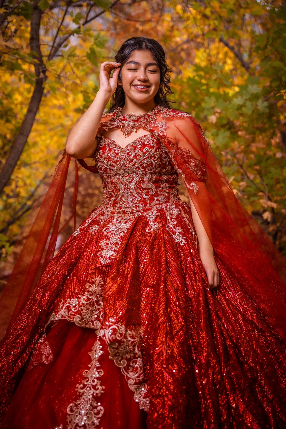 Smiling young woman in a red embroidered quinceañera ball gown posing among colorful autumn trees.