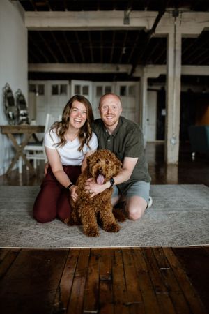 A couple poses with their fluffy brown dog on a rustic wooden floor in an industrial-style space.