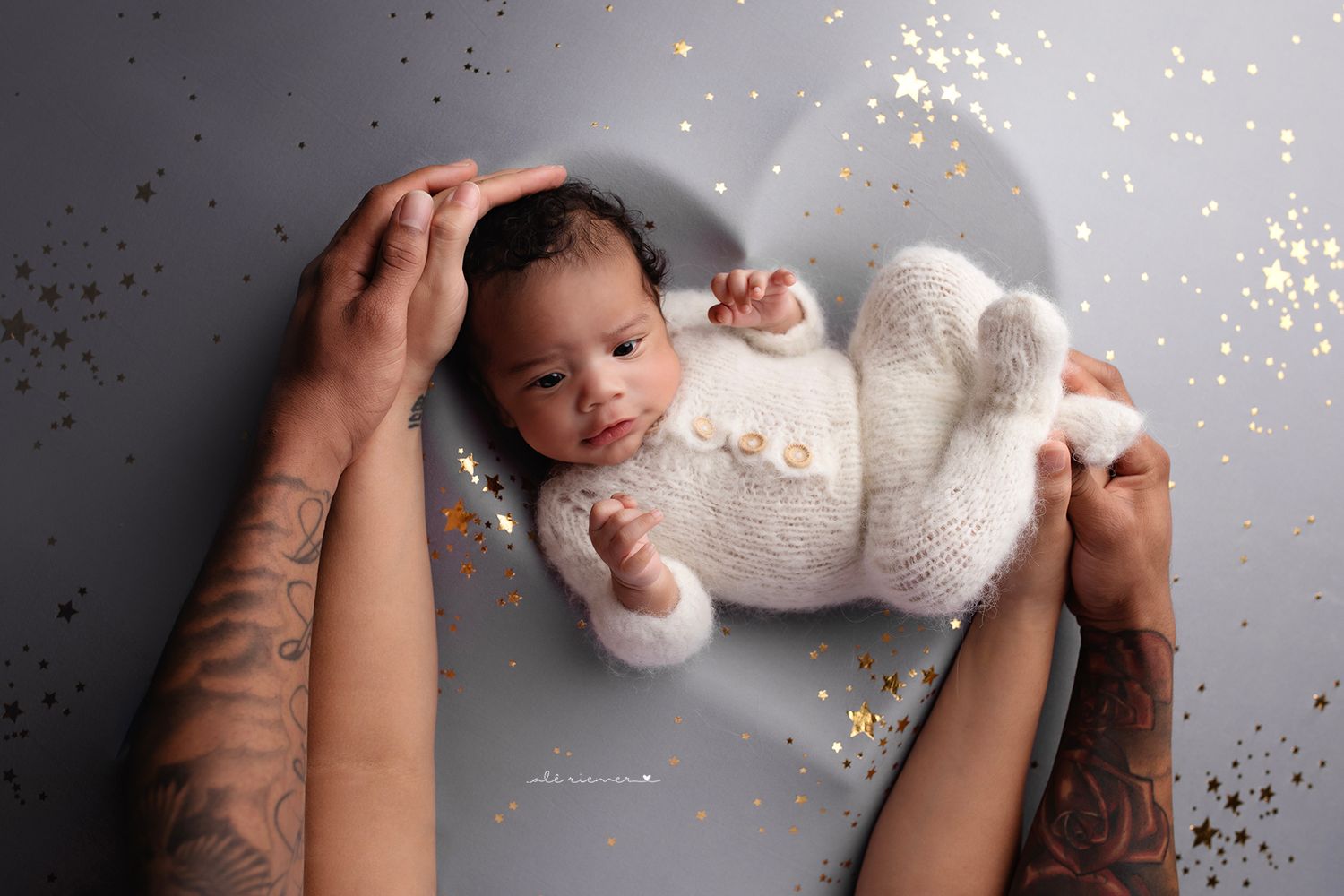 Newborn in white outfit surrounded by glitter while being held in tattooed hands.