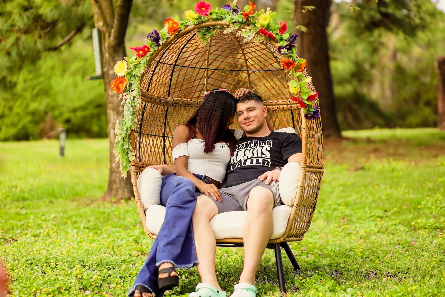 Couple sitting together in decorative wicker egg chair with colorful flowers outdoors.
