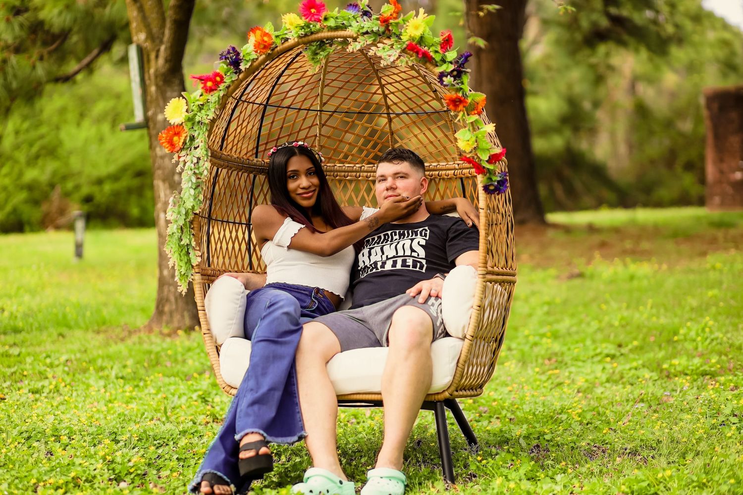 Interracial couple posing in floral decorated wicker egg chair on green lawn.