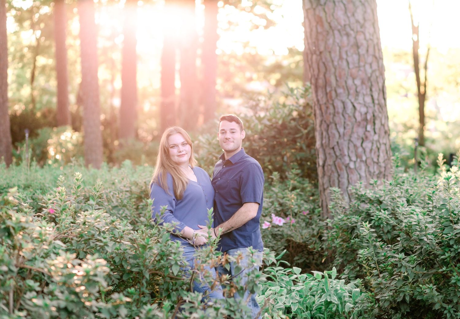Couple standing together among wildflowers in a sunlit forest during golden hour.