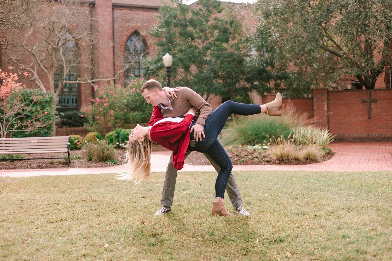 Couple dancing outdoors near a brick church, man dipping woman in red cardigan on green lawn.