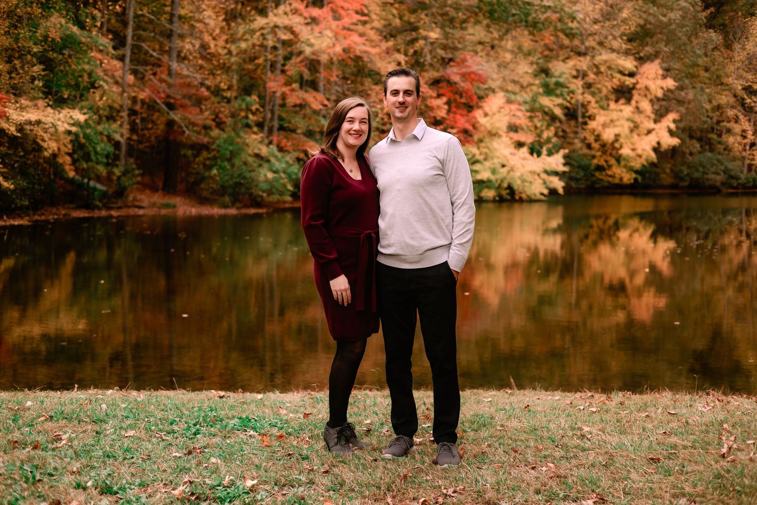 Couple smiling together beside a reflective pond surrounded by vibrant autumn foliage.