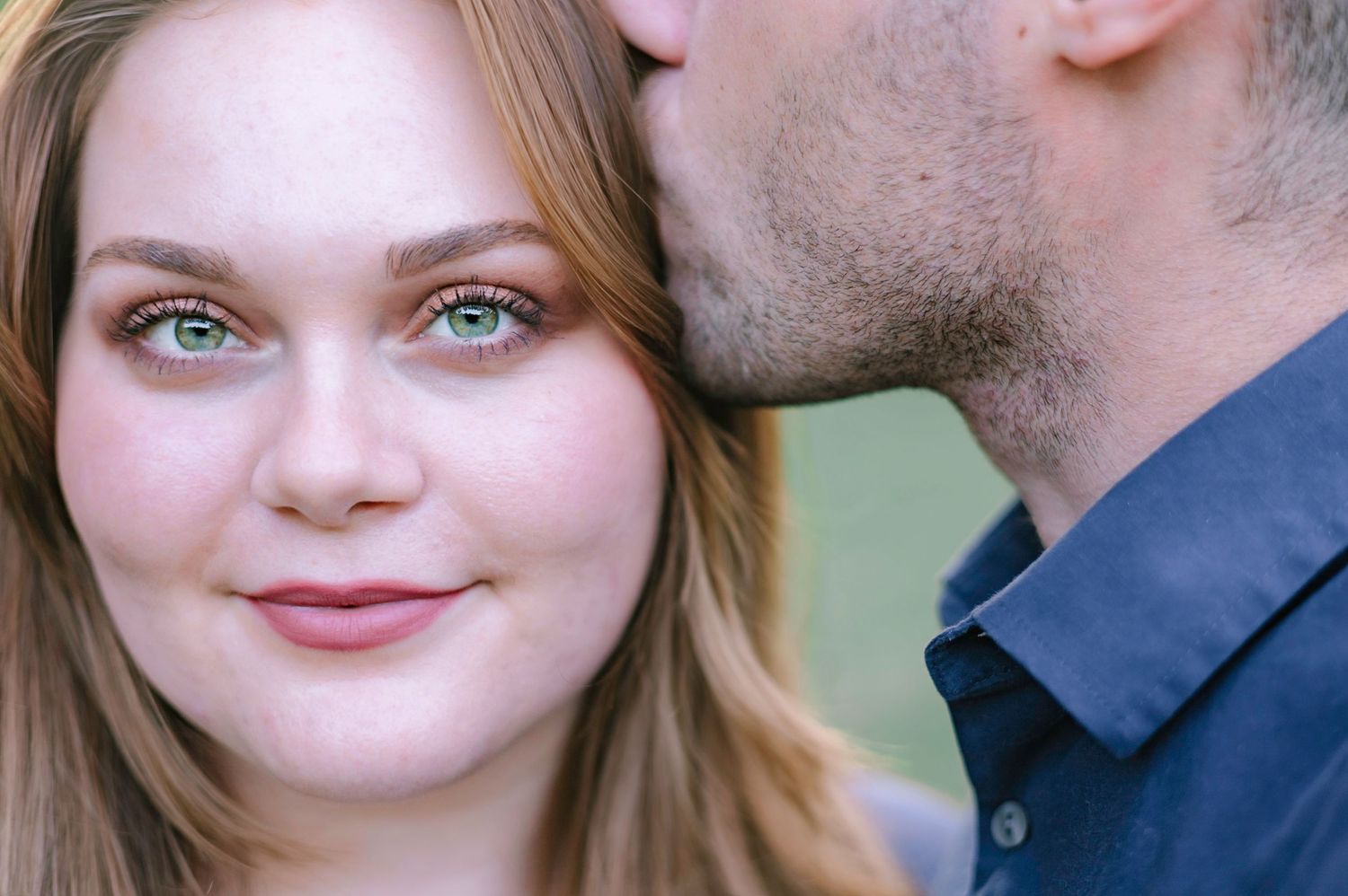 Close-up of a woman with green eyes smiling as her partner kisses her cheek outdoors.