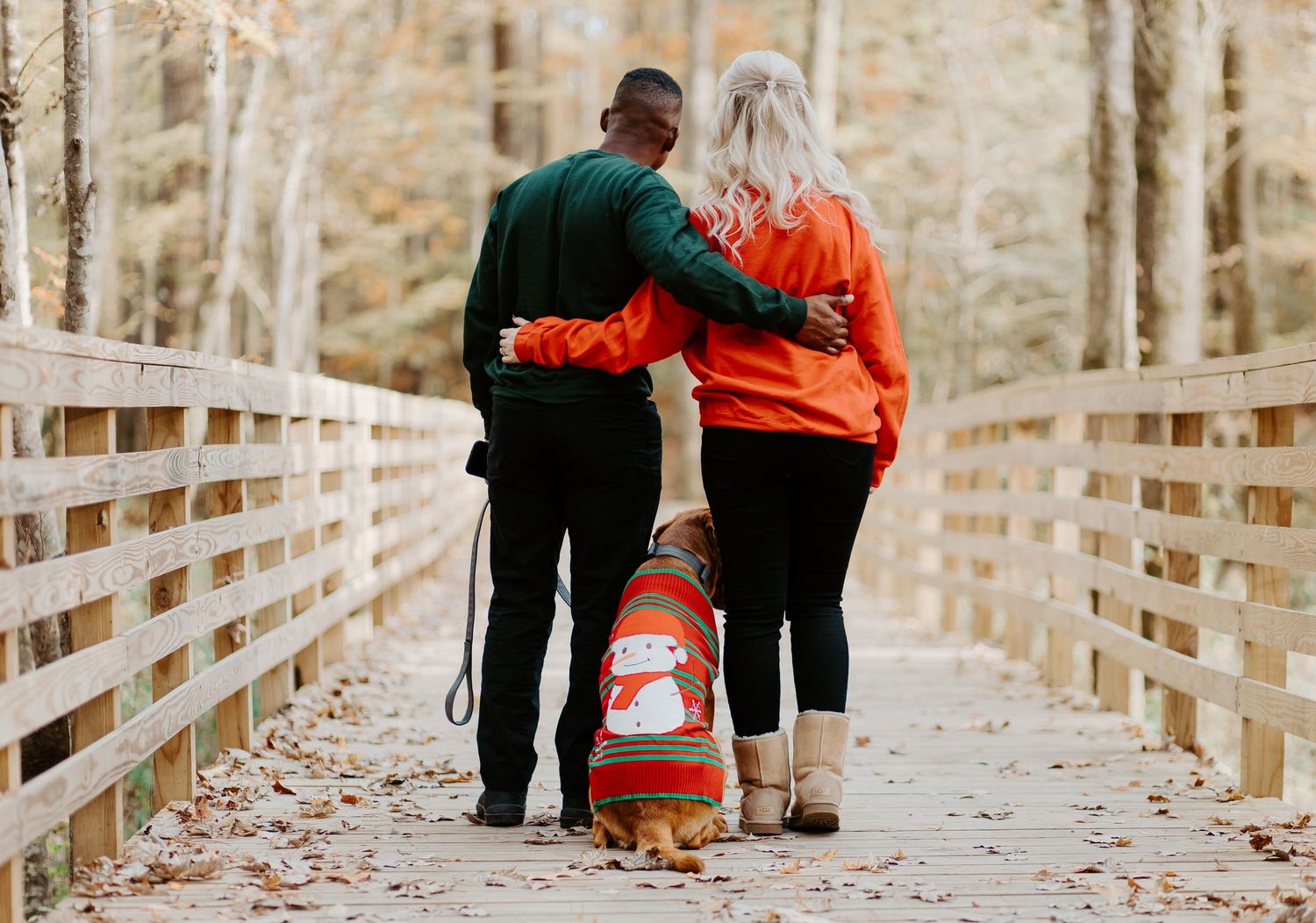 Couple walking their dog in a festive sweater on a leaf-covered wooden bridge.