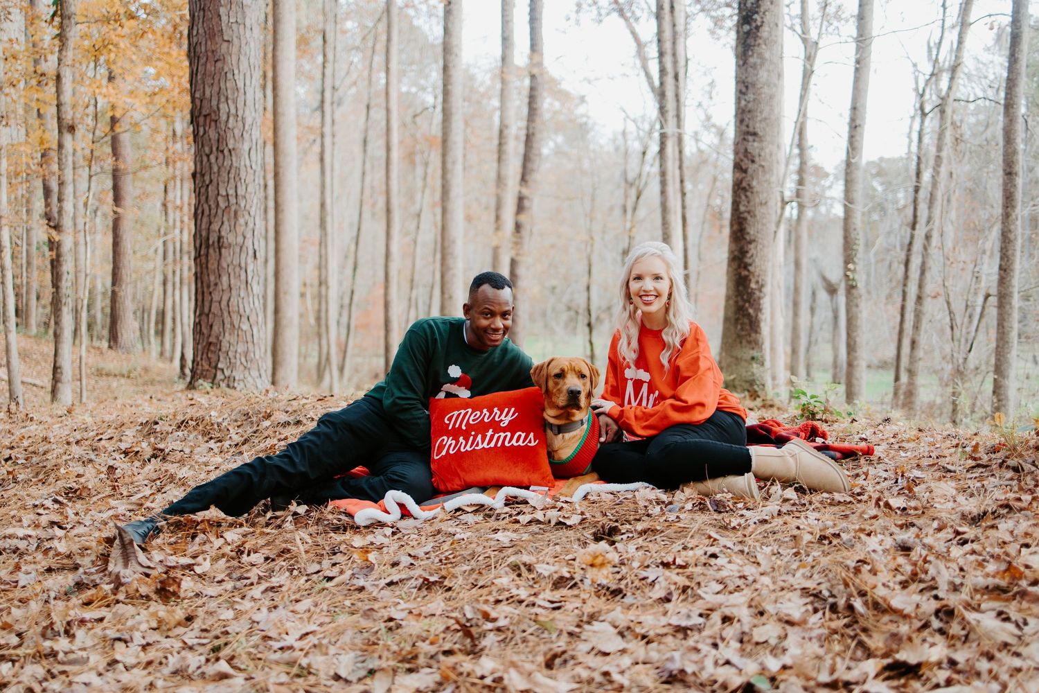 Couple sitting on fall leaves in woods with golden dog holding Merry Christmas pillow.