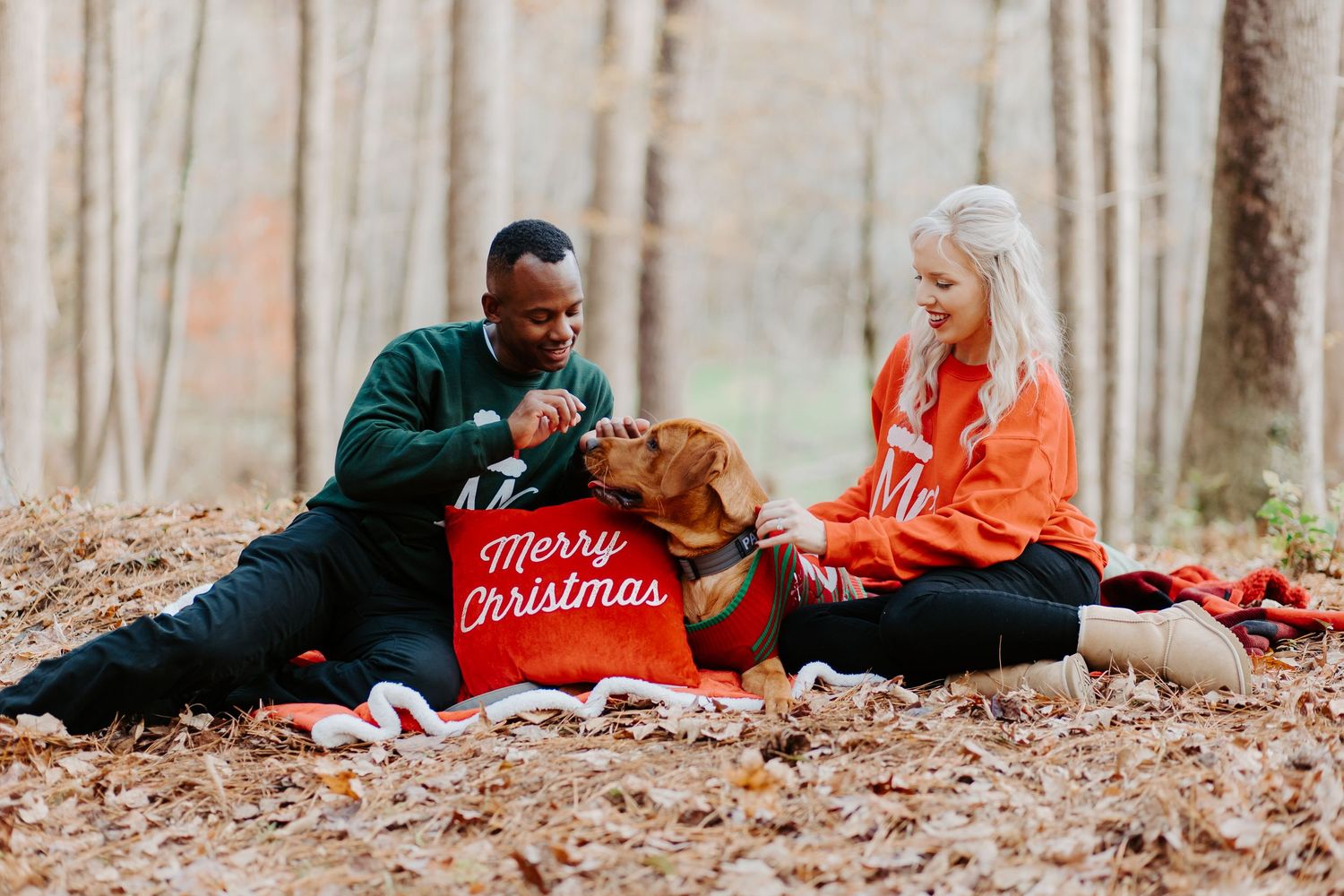 Man and woman laughing with golden retriever and Merry Christmas pillow in autumn forest.