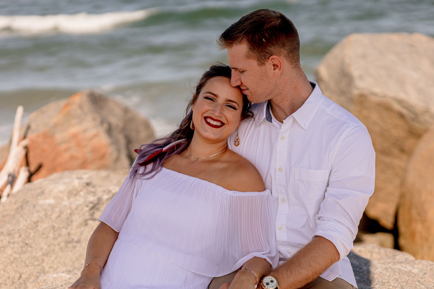 Happy couple sitting on beach rocks, woman in white dress laughing as man kisses her cheek.