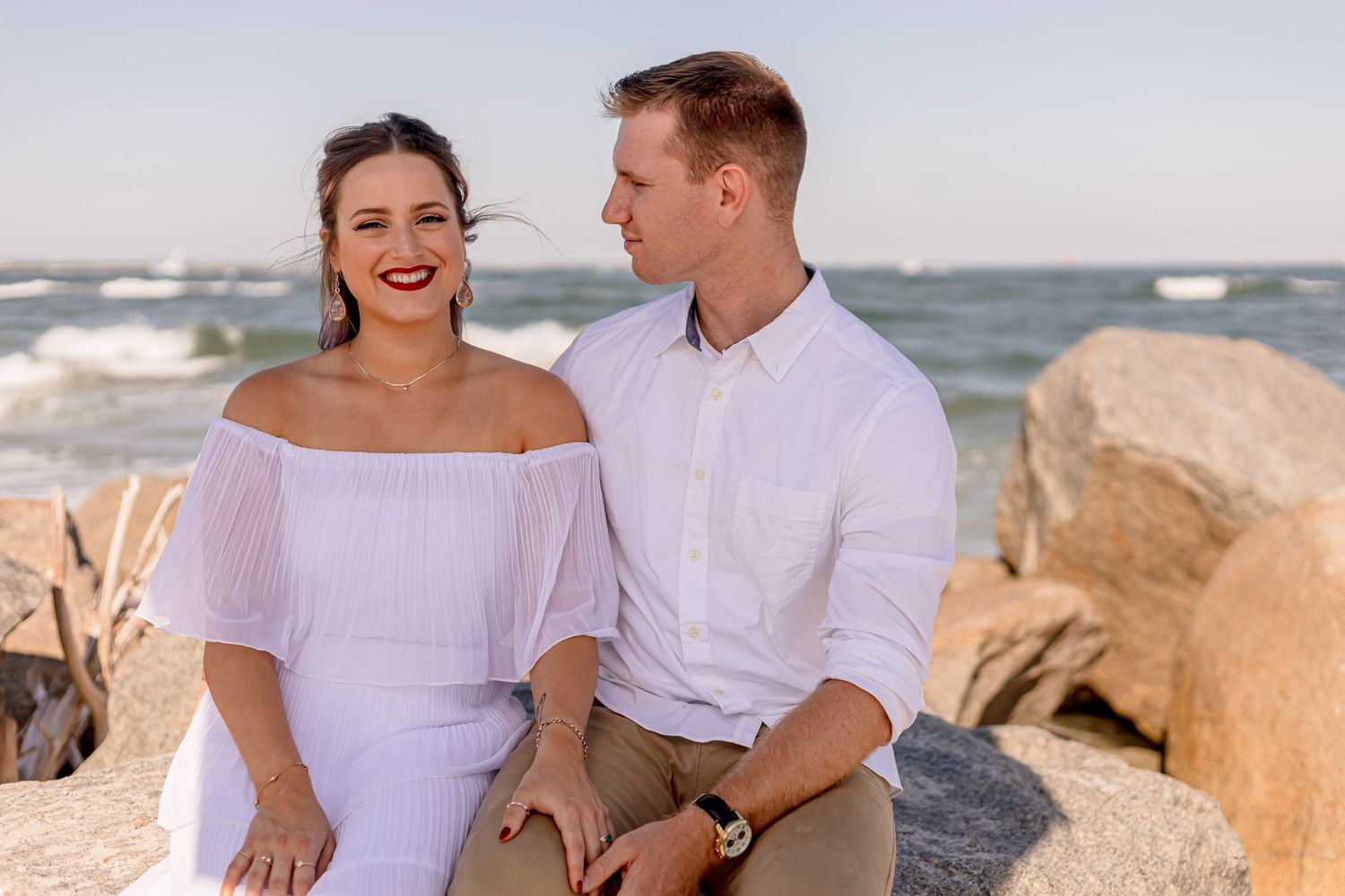 Couple sitting together on beach rocks, woman in white off-shoulder dress with red lips smiling.