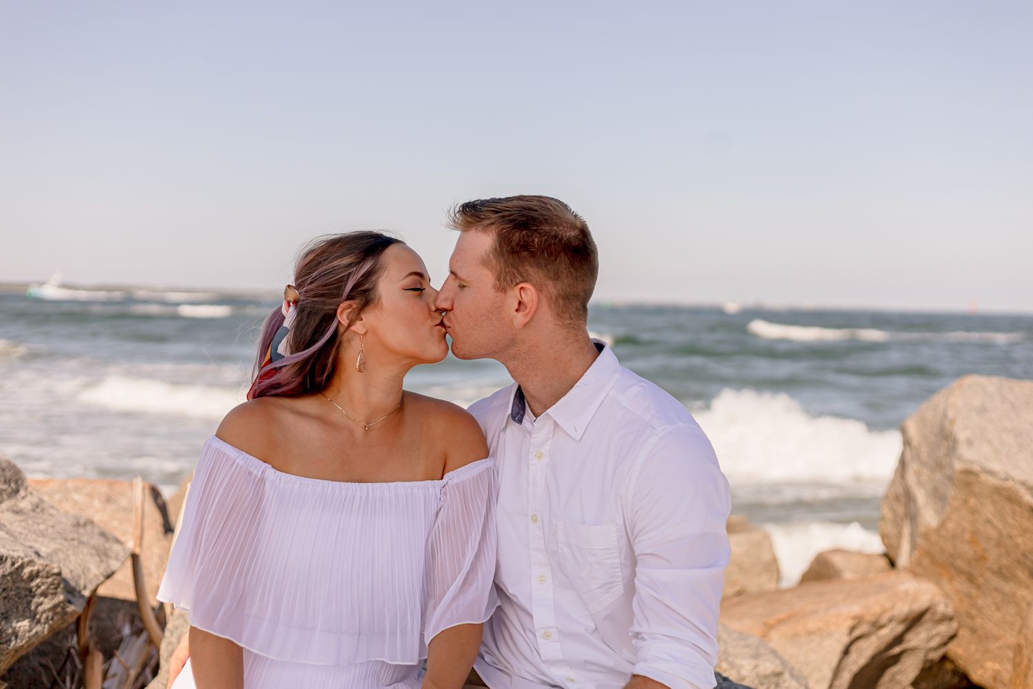Romantic couple sharing a kiss on rocky beach with ocean waves in the background.