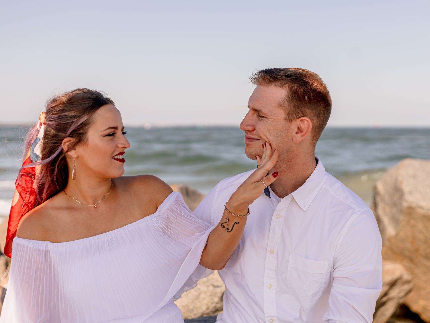 Smiling couple at the beach, woman in white off-shoulder dress gently touching mans face.