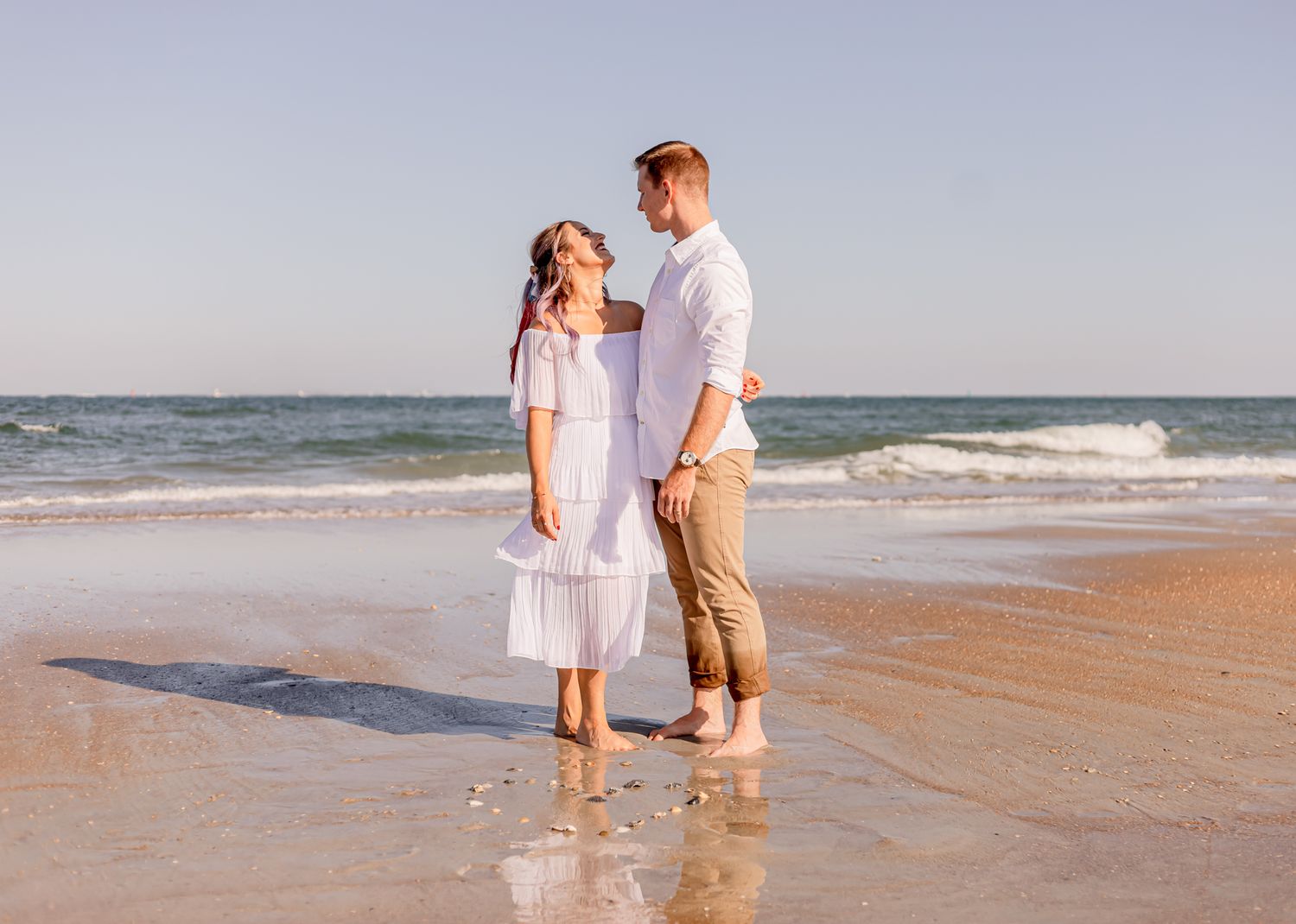 Young couple in white outfits smiling at each other while standing in ocean waves on beach.