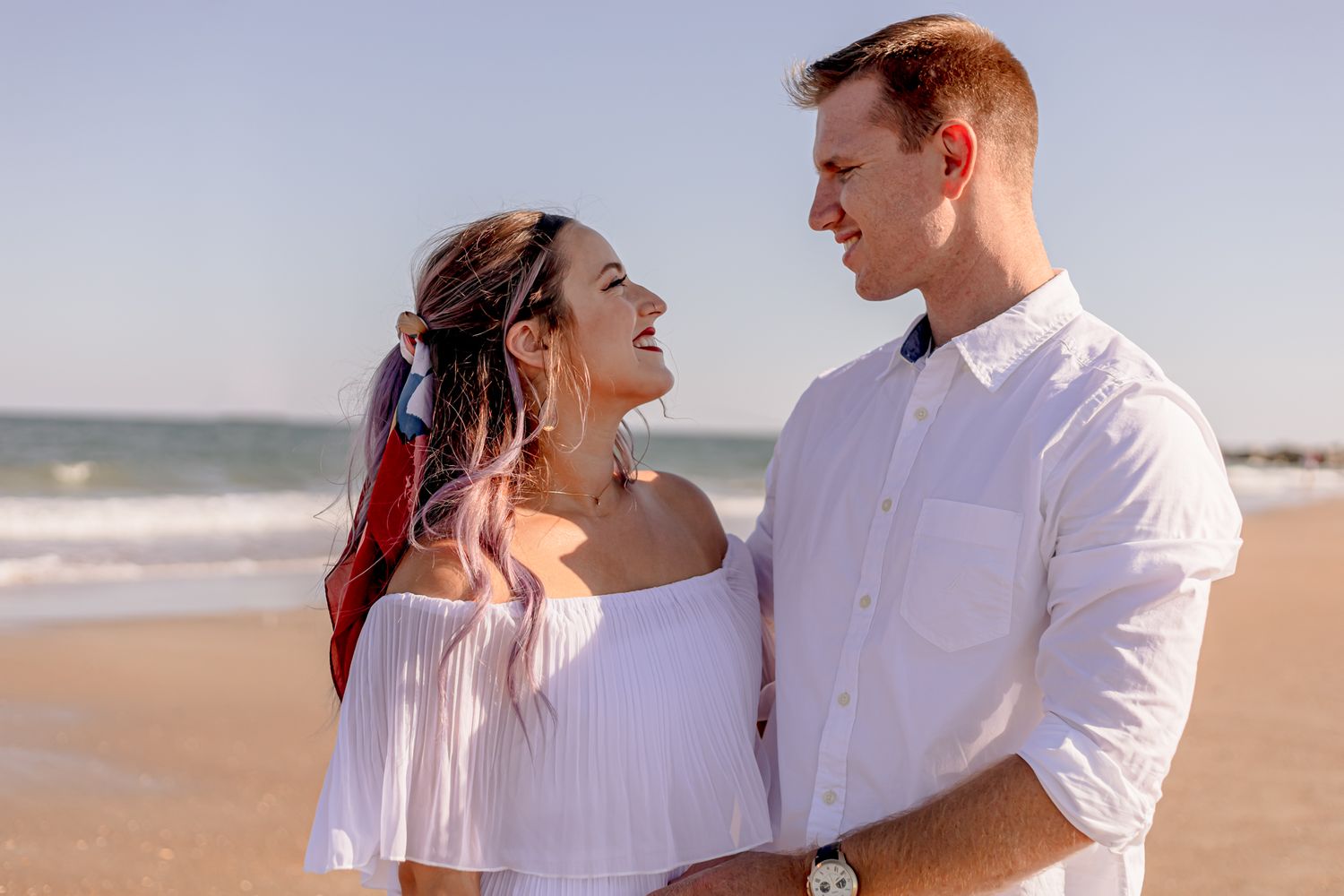 Happy couple laughing together on sunny beach, woman with colorful hair highlights in white dress.