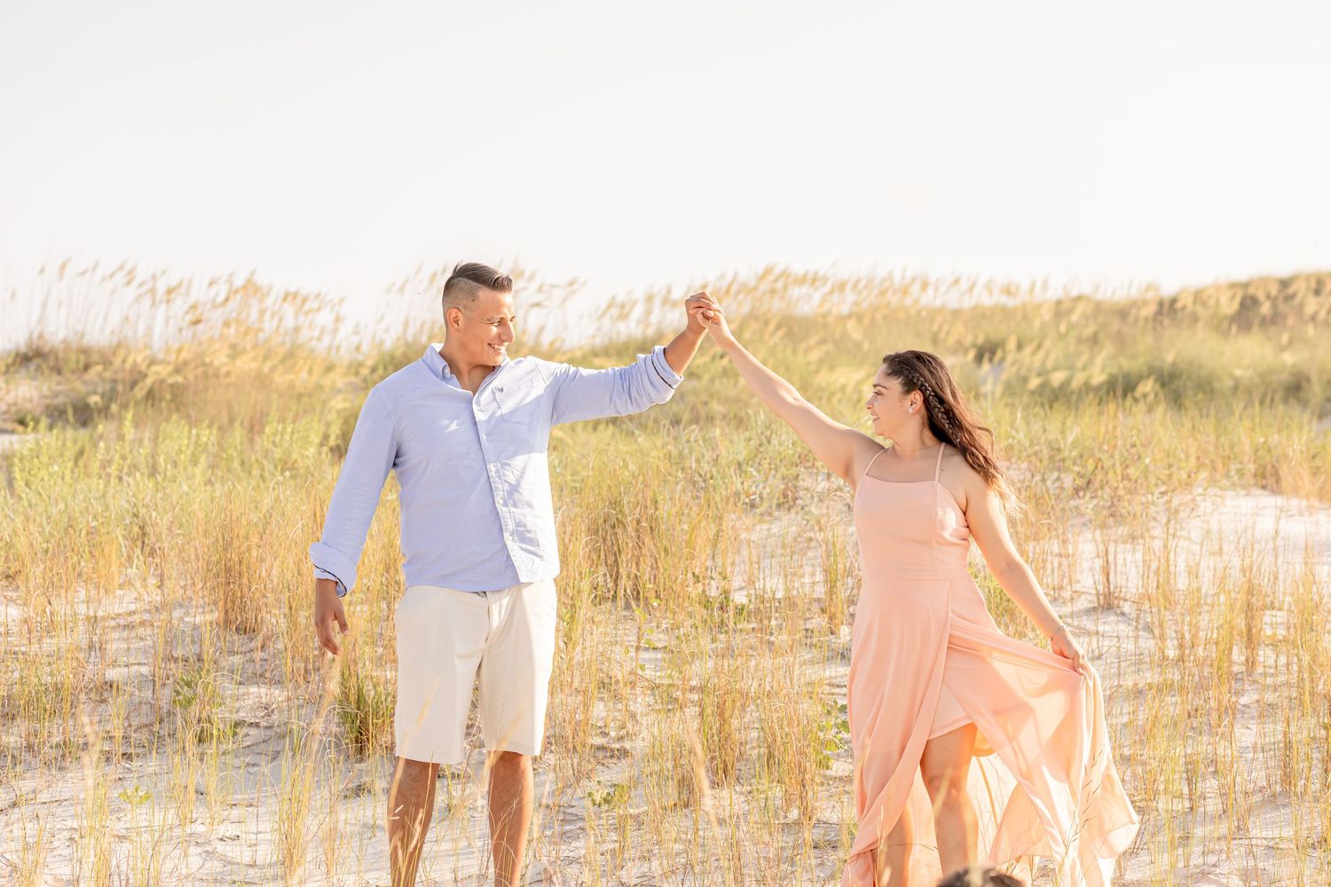 Joyful couple holding hands and spinning in flowing peach dress among beach dunes at sunset.