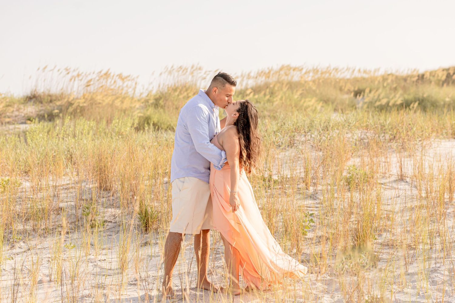 Romantic couple embracing and kissing among tall grass on sandy beach dunes at sunset.