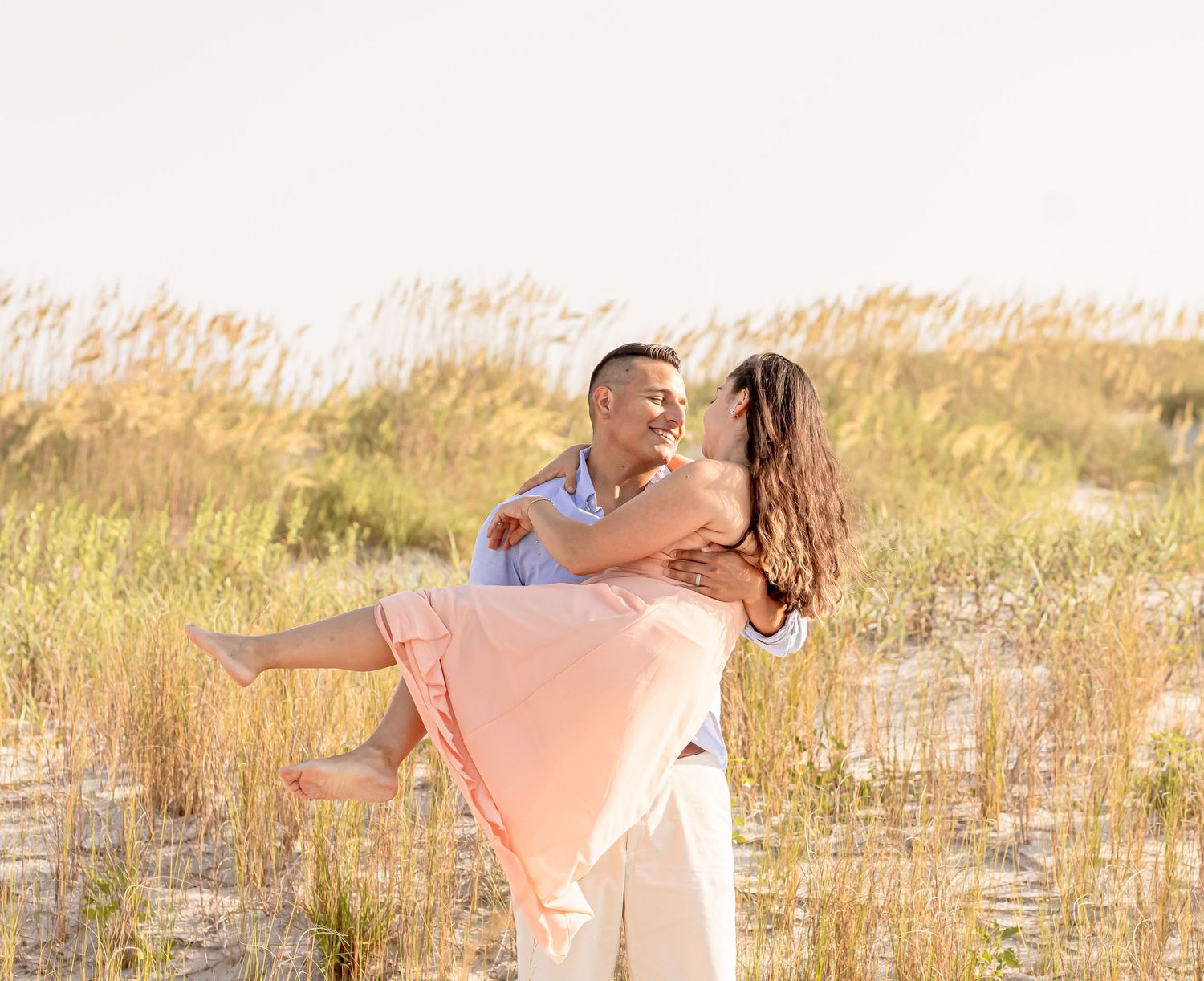 Man joyfully lifting woman in flowing peach dress among golden sea oats on beach.