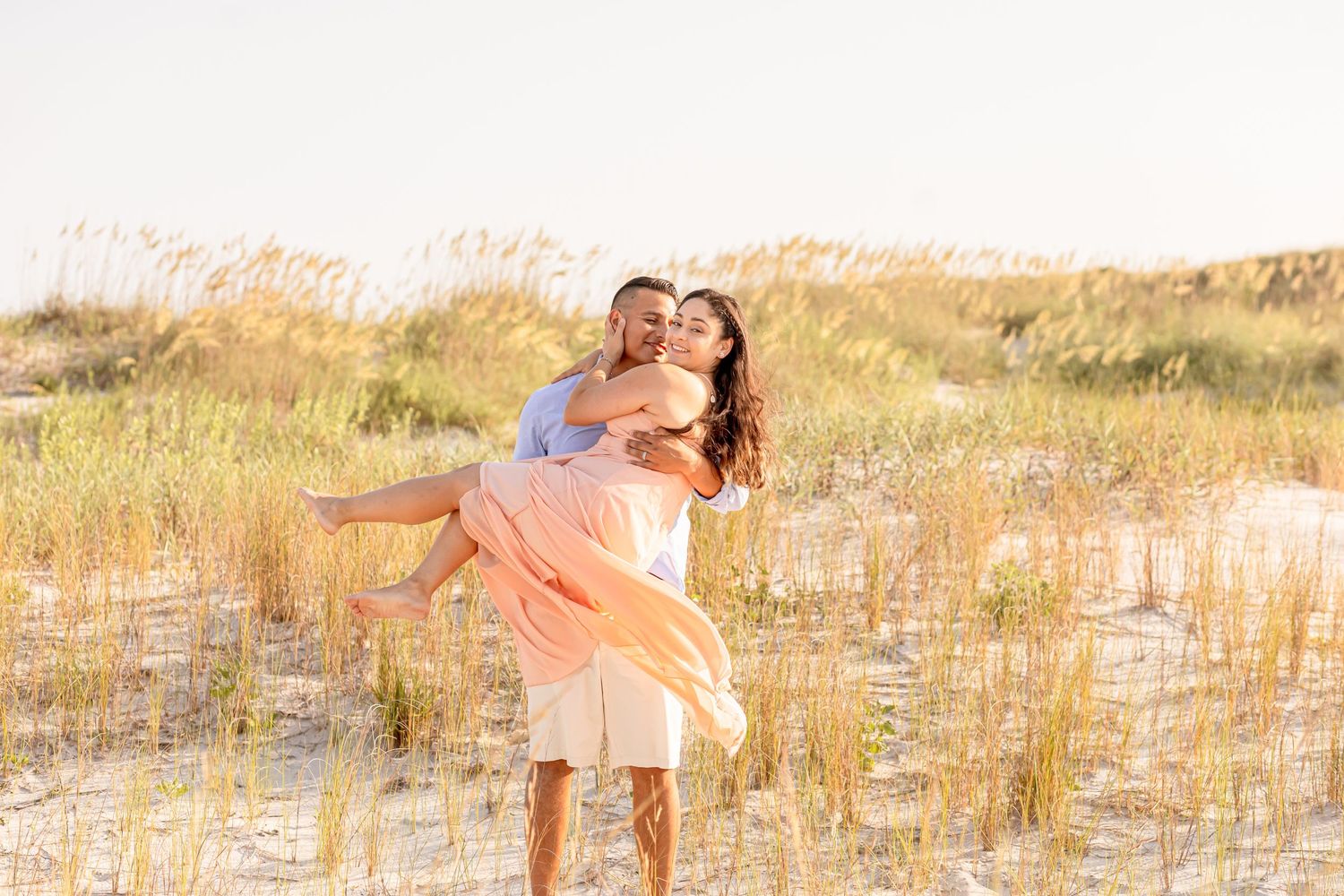 Man joyfully lifting woman in peach dress among sandy dunes and tall grass at golden hour.