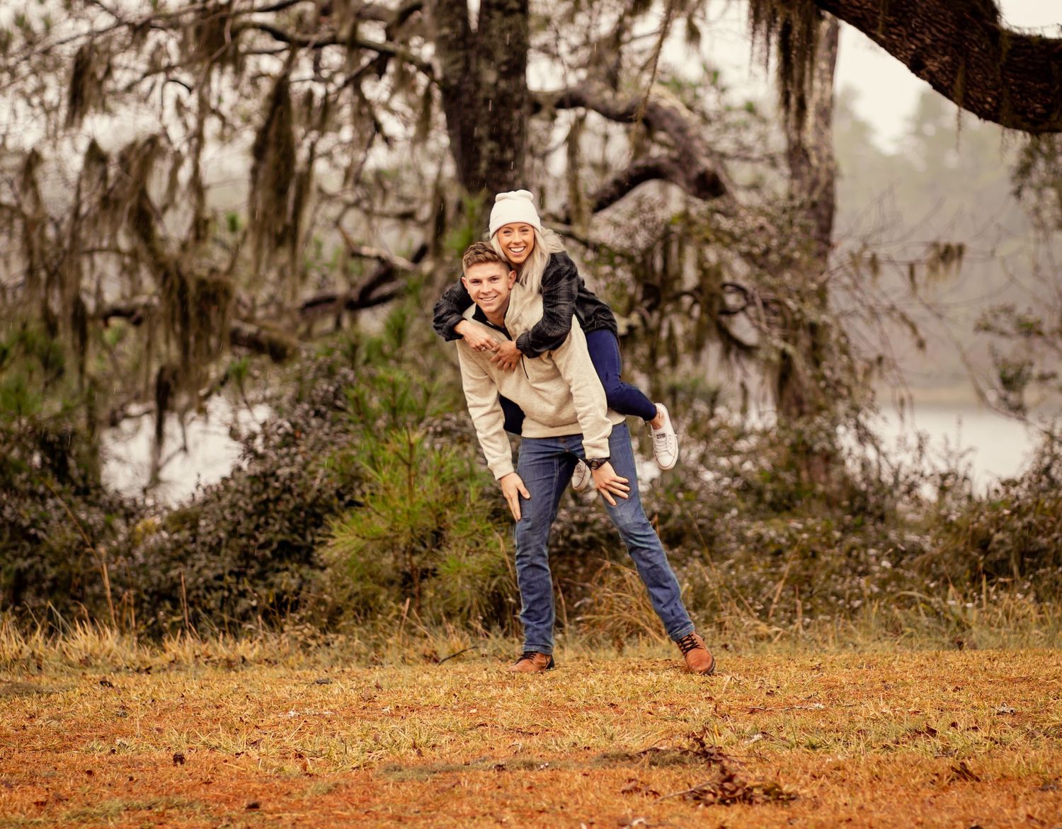 Couple laughing as man gives woman piggyback ride under Spanish moss tree.