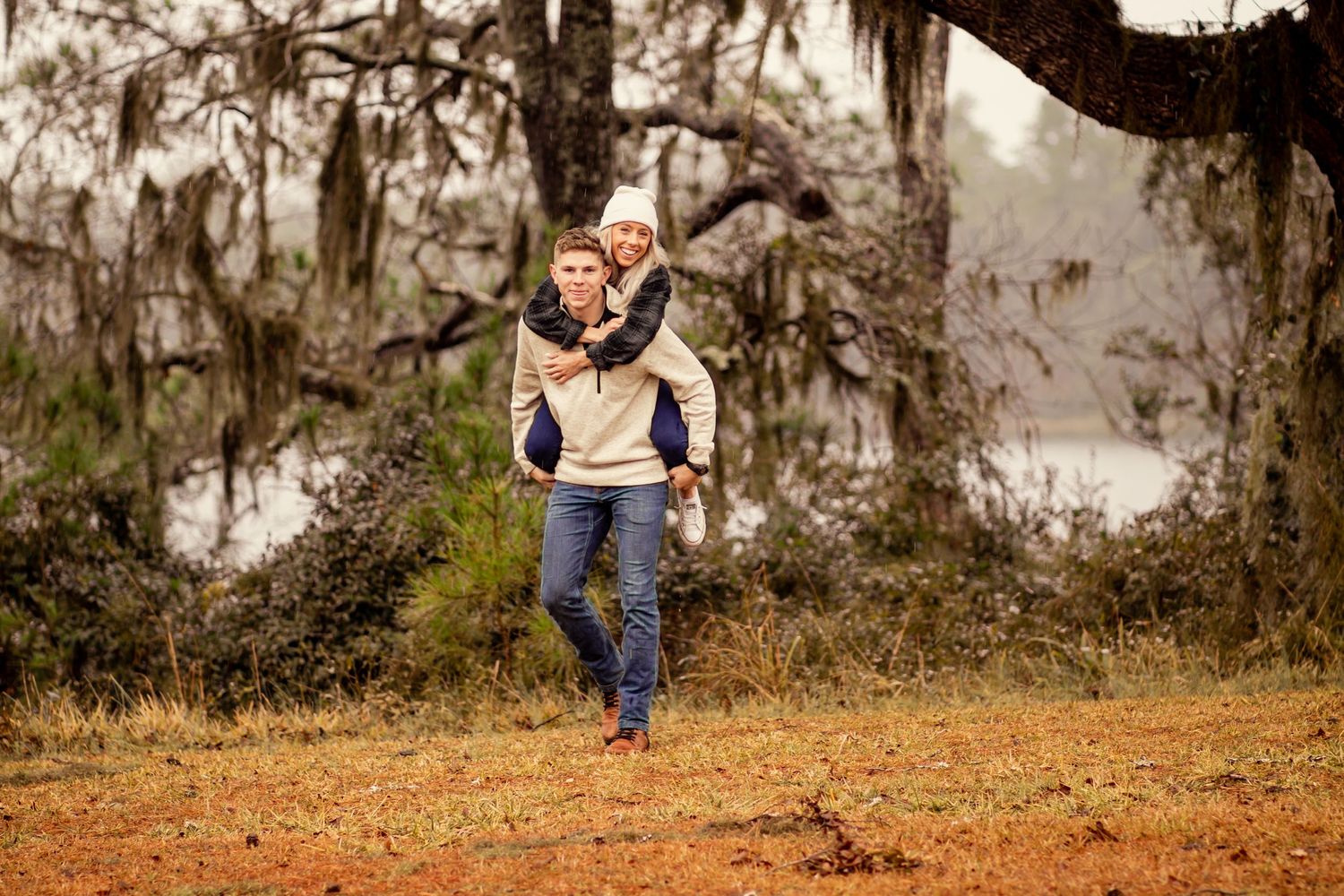 Man giving woman piggyback ride through rustic field with Spanish moss trees.