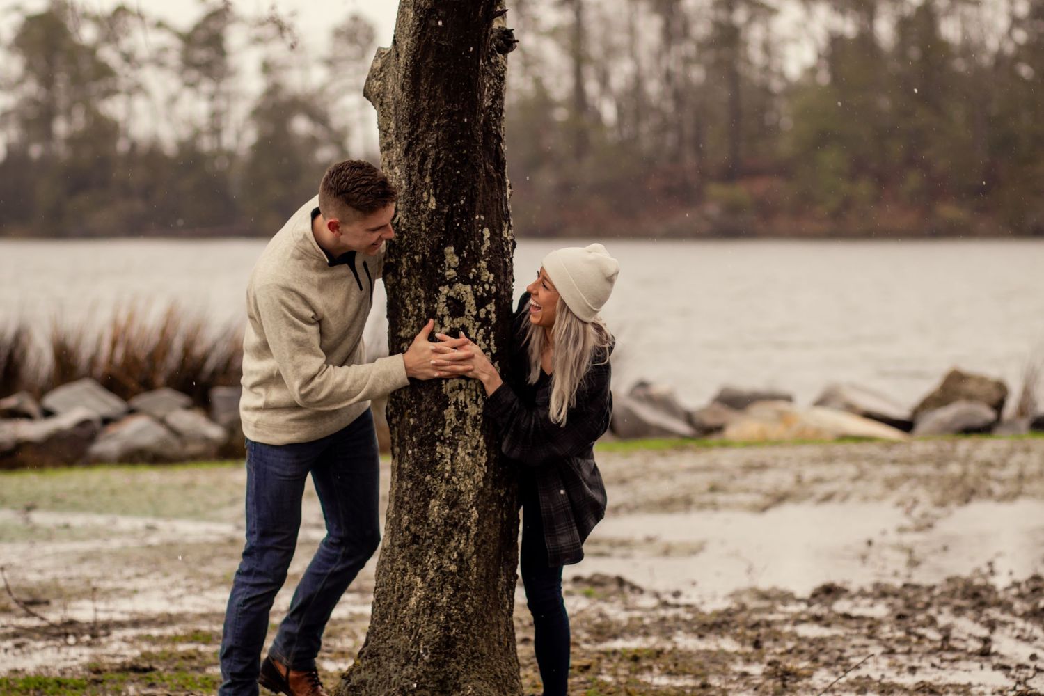 Playful couple peeking around tree trunk near rocky lakeside on cloudy day.