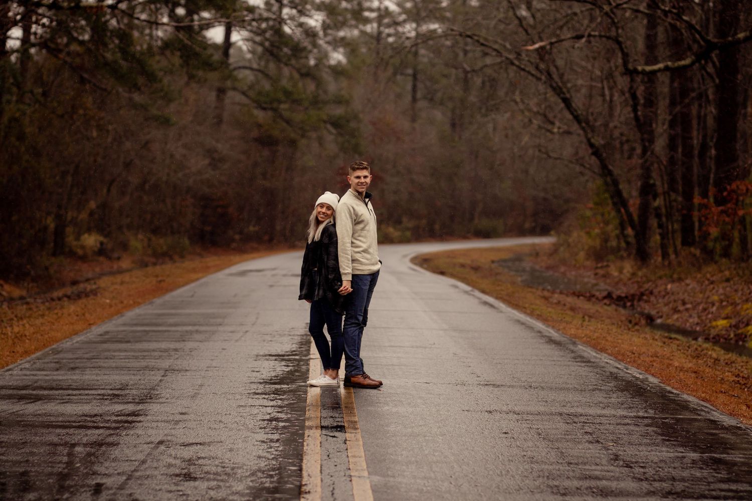 Couple standing together on a rain-soaked road surrounded by autumn trees.