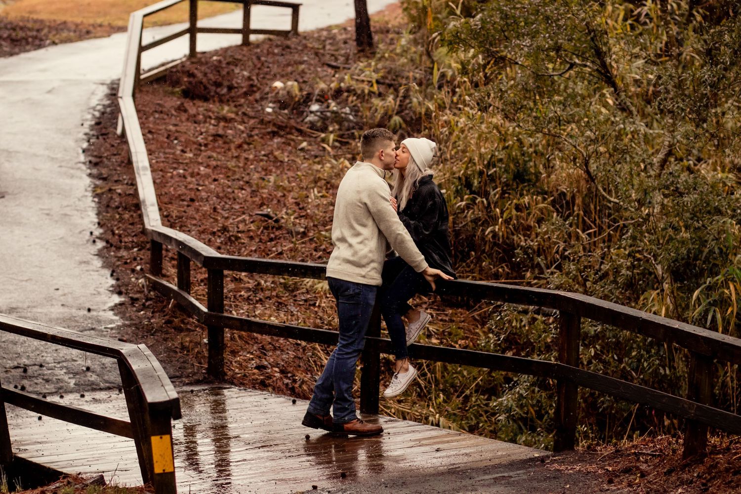 Couple sharing a kiss on a wet wooden bridge railing in the rain.